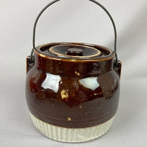 May include: A brown and white glazed ceramic crock with a wire handle and a lid. The crock has a white band around the bottom.