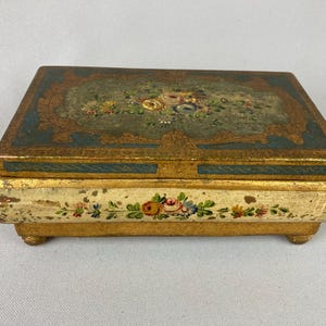 May include: A rectangular, antique jewelry box with a distressed, gold-painted finish. The lid features a teal background with floral artwork. The box's base is cream-colored with painted floral accents. The box is raised on small, rounded feet.