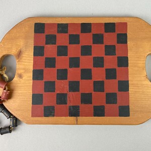 Op de afbeelding: Een ovaal houten dienblad met een natuurlijke lichtbruine afwerking en twee geïntegreerde handgrepen. Het midden is voorzien van een geschilderd dambordpatroon met afwisselend rode en zwarte vierkanten. Een snoer van houten kralen is bevestigd.