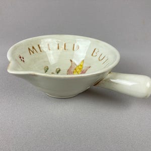 May include: Off-white ceramic butter warmer with a handle and a pouring spout. The words "MELTED BUTTER" are written in brown. The bowl is decorated with a corn and leaf design.