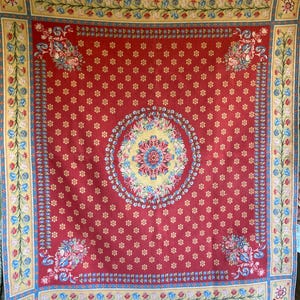 May include: A square textile featuring a deep red background with a repeating pattern of small yellow flowers. A large central circular medallion displays intricate floral motifs in yellow, blue, and pink. The border is decorated with blue and yellow floral vines.