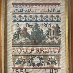 May include: A framed sampler with a white background and colorful embroidery. The sampler includes the alphabet, the date "12 Dec 1801", a scene with two houses and trees, and the year "1956".