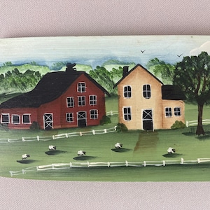 May include: A hand-painted wooden sign depicting a rural scene with two farmhouses, one red and one yellow, surrounded by green fields and white fences. There are white sheep in the field.