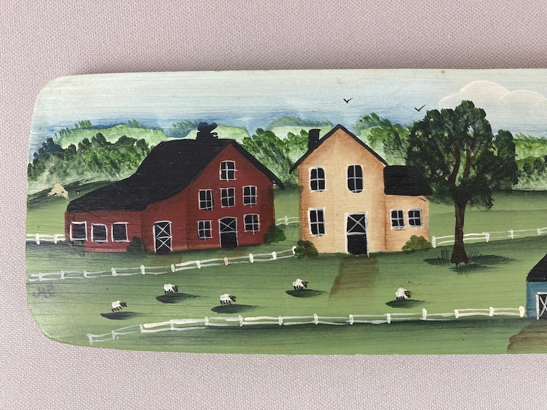 May include: A hand-painted wooden sign depicting a rural scene with two farmhouses, one red and one yellow, surrounded by green fields and white fences. There are white sheep in the field.