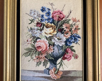 May include: A framed needlepoint embroidery depicting a bouquet of colorful flowers in a blue and white vase. The flowers include pink, white, yellow, and blue blooms.