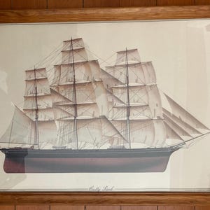 May include: Framed print of the 'Cutty Sark' clipper ship. The ship is depicted with full sails, set against a neutral background. The hull is a deep burgundy, and the frame is a light brown wood.