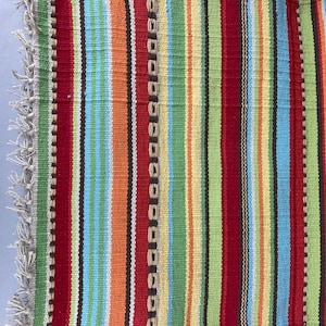 May include: A colorful, striped woven textile with a fringed edge. The design features vertical stripes in red, orange, green, yellow, blue, and black. The textile is likely a rug or decorative fabric.