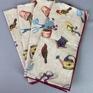 May include: Set of five beige linen napkins with a red border. The napkins feature a repeating pattern of gardening-themed illustrations, including birds, flowers, watering cans, and gardening tools.