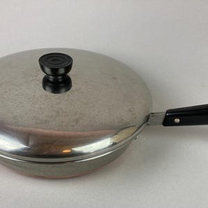May include: Stainless steel frying pan with a lid and a black handle. The pan has a round shape and a silver finish. The handle is attached with rivets and has a loop at the end.