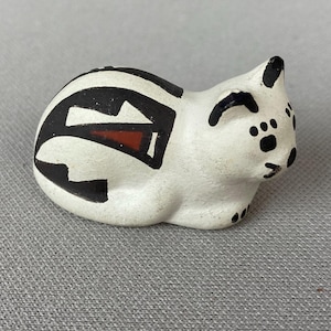 May include: A small, white ceramic cat figurine with black and red geometric designs. The cat is in a resting pose, with black spots on its face and paws. The figurine is a decorative item.