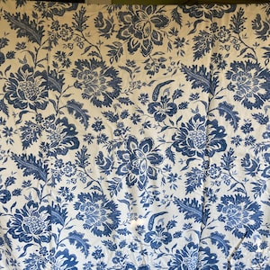 May include: A fabric with a repeating floral pattern in shades of blue on a cream background. The design features large, stylized flowers, leaves, and smaller floral accents, creating a classic, decorative textile.