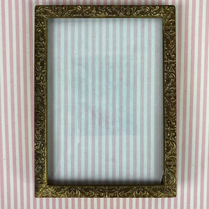 May include: Rectangular antique gold picture frame with ornate floral detailing. The frame is empty, with a clear glass insert. The background is a pink and white striped pattern.