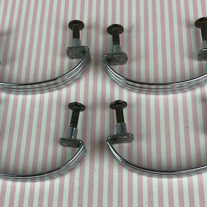 May include: Four silver-toned metal drawer pulls with a curved, semi-circular design. Each handle features two vertical mounting posts with threaded screws attached, showcasing a polished finish and a simple, clean structure for furniture hardware.