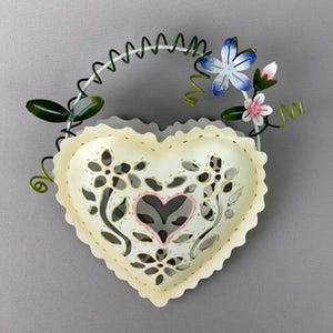 May include: A cream-colored, heart-shaped metal wall hanging with floral cutouts and a pink heart in the center. It features a white handle with green leaves and vines, and blue and pink flowers. The decorative piece is designed for wall display.