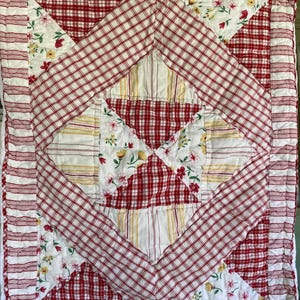 May include: A handmade quilt featuring a patchwork design with red and white gingham, floral, and striped patterns. The quilt has a diamond-shaped center and a red and white striped border. The color palette includes red, white, and yellow.