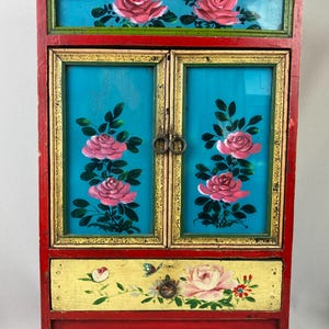 May include: A small, hand-painted cabinet with a red frame and a yellow drawer. The cabinet doors and top section feature a turquoise background with pink rose floral designs. The drawer is decorated with a rose, butterfly, and other floral elements.