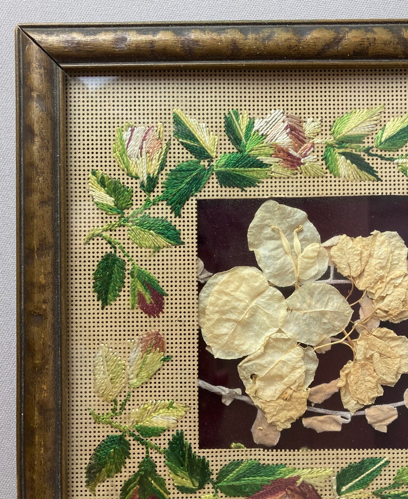 Op de afbeelding: Een ingelijst borduurwerk met een bloemenmotief. De borduurwerk is uitgevoerd in tinten groen, wit en bruin. De lijst is van hout en heeft een gouden afwerking.