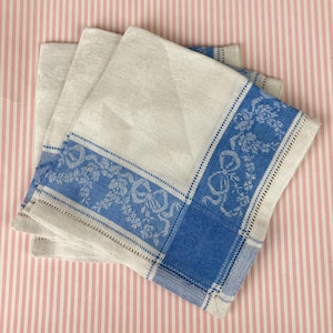May include: A stack of white linen napkins with a blue border featuring floral and bow designs. The napkins are folded and stacked, showcasing the detailed pattern and texture of the fabric. The background is a pink and white striped pattern.