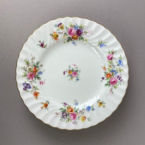 May include: A white porcelain plate with a gold rim and a floral pattern in shades of pink, yellow, blue, and green.