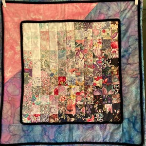May include: A square patchwork quilt featuring a central grid of small floral fabric squares that transition from light pastel tones to dark, vibrant colors. The quilt is framed by a black border and surrounded by a pink and blue tie-dye fabric.