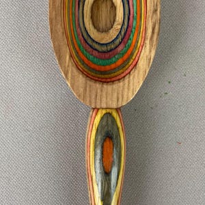 May include: Wooden spoon with a colorful, concentric circle design in the bowl. The spoon's bowl features rings of orange, green, yellow, and blue. The handle has a similar layered design with red, yellow, and orange tones. A unique kitchen utensil.