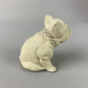 Pu&ograve; includere: Una piccola statuetta di ceramica bianca di un bulldog francese seduto con la testa girata di lato. La statuetta ha un collare di perline.