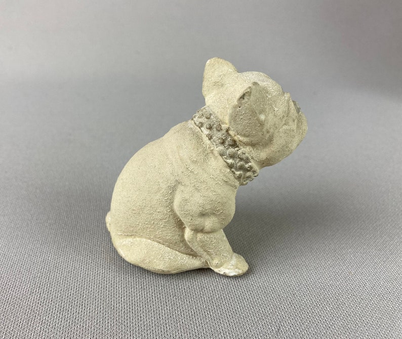 Pu&ograve; includere: Una piccola statuetta di ceramica bianca di un bulldog francese seduto con la testa girata di lato. La statuetta ha un collare di perline.