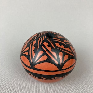 May include: A small, round, terracotta pot with a black geometric design. The pot has a small opening at the top.