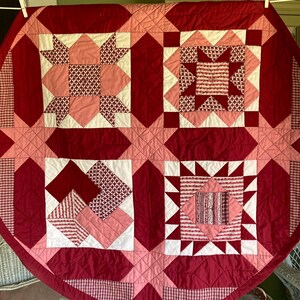 May include: A handmade quilt with a geometric pattern in shades of red, pink, and white. The quilt features star and square designs, with a diamond-stitched background. The overall design is symmetrical and visually appealing.