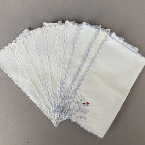 May include: A collection of white handkerchiefs with scalloped edges and delicate blue stitching. Each handkerchief features a small embroidered floral design with red accents. The handkerchiefs are neatly arranged, fanned out against a neutral background.