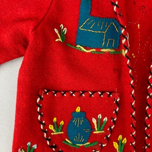 以下が含まれることがあります： 赤いウールジャケットに刺繍が施されています。ジャケットには青い家と青い花瓶に黄色い花が刺繍されています。すべてカラフルな糸で刺繍されています。