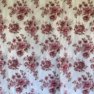 May include: A white fabric featuring a repeating pattern of dusty rose floral bouquets. The design consists of clusters of roses and smaller blossoms in shades of muted pink and mauve, evenly distributed across the entire surface of the textile.