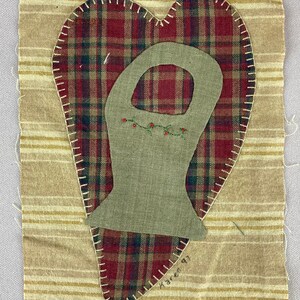 May include: A heart-shaped quilted wall hanging with a red and green plaid fabric background. A green fabric cutout of a boot with a vine and red berries embroidered on it is centered on the heart. The piece is signed "Karen 97" in the bottom right corner.
