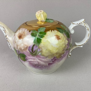 May include: A porcelain teapot with a gold lid and a floral design. The teapot features a white handle and spout with gold trim. The body of the teapot is decorated with large, colorful flowers in shades of yellow, purple, and white.