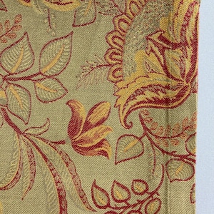 May include: Close-up of a decorative textile with a floral pattern. The fabric features a repeating design of stylized flowers and leaves in shades of gold and red against a muted green background. The texture of the fabric is visible.