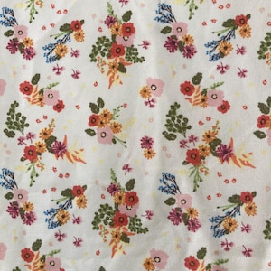 May include: White fabric with a repeating floral pattern. The design features clusters of flowers in shades of red, orange, pink, and purple, with green leaves and small blue accents. The background is a soft white.