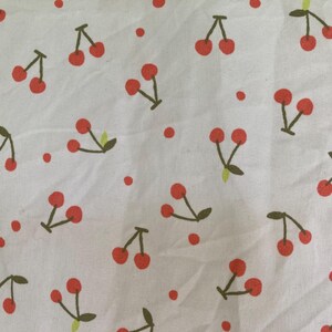 May include: White fabric with a repeating pattern of red cherries and green stems. The cherries are in pairs and singles, with small red dots scattered throughout the design. This fabric is suitable for various sewing projects.