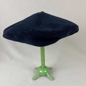 May include: A navy blue beret sits atop a light green display stand. The beret is made of a soft, textured material. The stand has a round base with three legs and a vertical pole.