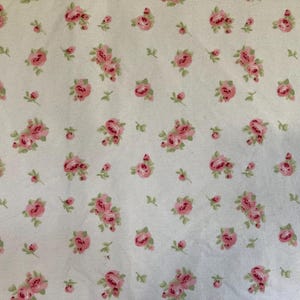May include: Cream-colored fabric with a repeating pattern of pink roses and green leaves. The roses vary in size and are clustered together, creating a delicate floral design. The fabric has a soft, vintage aesthetic.