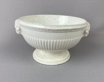 May include: A white ceramic bowl with a pedestal base. The bowl has a fluted body and decorative rope-like trim around the rim and base. Two ornate handles are on either side of the bowl.