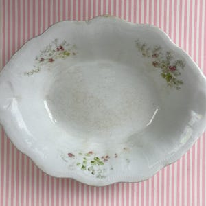 May include: A white, antique oval serving dish with a scalloped edge. The dish features delicate floral accents in pink and green. The dish is likely made of porcelain or ceramic and is a vintage piece.