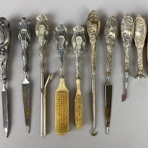 May include: A collection of antique silver-toned tools, including knives, a hook, and a brush. The handles feature ornate floral and scroll designs. Some tools have ivory or bone handles. The tools are arranged on a light gray surface.