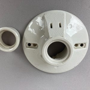 May include: A white ceramic light fixture base and a separate socket ring. The base has a circular design with a central opening and two screw holes. The socket ring is a smaller, cylindrical piece.