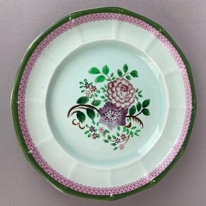 May include: A white ceramic plate with a scalloped edge, featuring a floral design in shades of purple, pink, and green. The rim is bordered with a pink and green pattern. The plate has a light blue center.