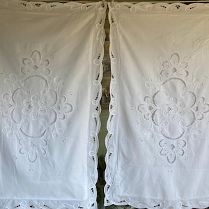 May include: Two white curtains with intricate floral embroidery. The curtains feature a scalloped edge and a large, symmetrical floral design in the center. The fabric appears to be a lightweight material.