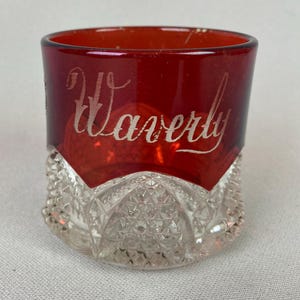 May include: A ruby red glass with the word "Waverly" etched in cursive. The top portion of the glass is red, while the bottom is clear with a diamond-cut pattern. A vintage-style drinking glass.