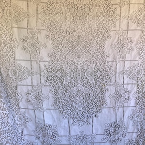 May include: A white lace tablecloth featuring an intricate floral pattern with repeating medallions and a grid-like structure. The fabric displays a delicate openwork design with detailed botanical motifs and symmetrical scrollwork throughout the textile.