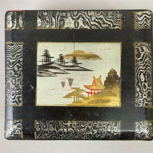 May include: A black wooden box with a silver and black patterned border. The box has a raised, rectangular panel with a painted scene of a Japanese garden with trees, a pagoda, and a lake.