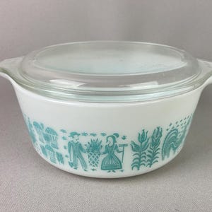 May include: White Pyrex bowl with a clear glass lid. The bowl features a turquoise design of people, plants, and animals around the exterior. The bowl has two handles and is on a gray surface.