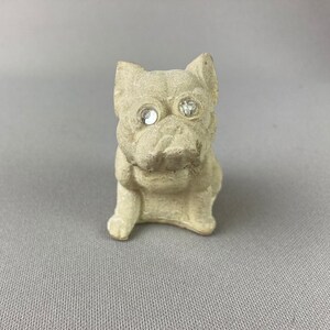 Pu&ograve; includere: Una piccola statuetta di ceramica bianca di un cane seduto con la testa leggermente inclinata di lato. Il cane ha due occhi di strass trasparenti.
